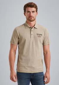 Béžová bavlněná polo tričko s límečkem, třemi knoflíky a potištěným textem na levé hrudi. Na sobě má modré džínové kalhoty.