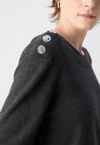 Un pull gris en matériau doux, avec un col rond et trois boutons décoratifs à l'épaule avec une finition texturée.