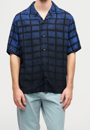 Mand iført kortærmet skjorte med blå og sort ternet mønster og lyseblå jeans, stående mod en ensfarvet baggrund.