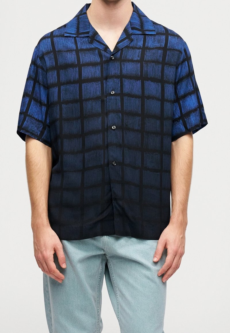 Homme portant une chemise à manches courtes à motif à carreaux bleu et noir et un jean bleu clair, debout devant un fond uni.