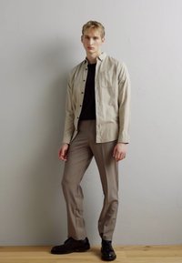 Camicia button-up beige chiaro sopra una maglietta nera, abbinata a pantaloni beige e scarpe nere, in piedi davanti a una parete neutra.