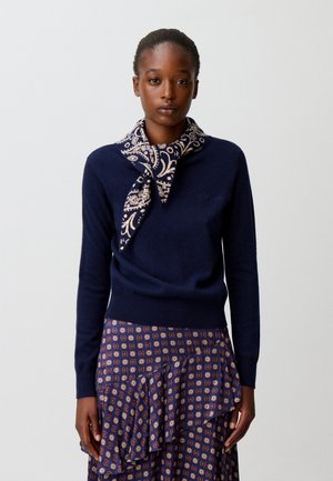 Mujer con suéter azul marino, bufanda con estampado de paisley y falda azul marino a juego con motivos circulares y detalle de volantes.