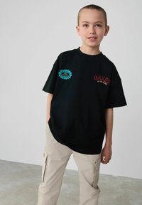Camiseta negra de manga corta con el texto "BAKER" en rojo y un parche gráfico en turquesa. Combinada con pantalones cargo beige, que presentan un ajuste relajado.