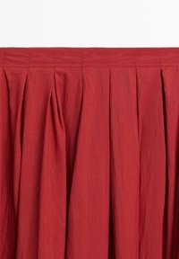 Punainen laskostettu hame, joka on valmistettu teksturoidusta kankaasta. Sisältää istuvan vyötärön, jossa on näkyvät ompeleet, sekä virtaavat laskokset, jotka laskeutuvat alaspäin.