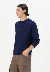 Hanorac bleumarin din bumbac, cu un guler rotund, manșete cu nervuri și un logo brodat de culoare aurie pe piept. Modelul poartă pantaloni cargo maro.