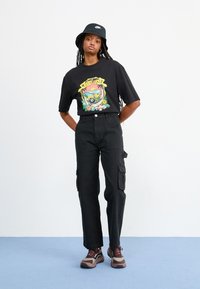 T-shirt grafico nero con design colorato, pantaloni cargo neri con tasche laterali, cappello a secchiello nero e scarpe da ginnastica marroni/grigie con dettagli rossi.