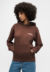 Femme aux cheveux bouclés portant un sweat à capuche marron ellesse, les mains dans la poche avant, et un jean bleu, regardant vers sa gauche.