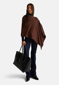 Hnědý vlněný pončo s drapovaným designem, spárovaný s tmavými riflovými kalhotami s rozšířenými nohavicemi a velkou černou koženou kabelkou. Hladká textura a strukturovaný tvar.