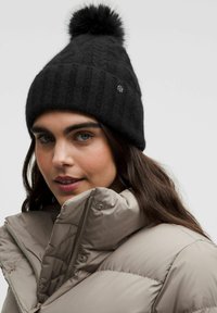 Gorro de punto negro con un patrón texturizado y pompón en la parte superior. Combinado con una chaqueta acolchada gris claro que presenta un cuello alto y cremallera.