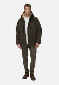 Parka-Jacke in Dunkelgrün mit einer hellbraunen, fleecegefütterten Kapuze, mehreren Fronttaschen und Reißverschlussdetails, kombiniert mit grauen Hosen und braunen Stiefeln.