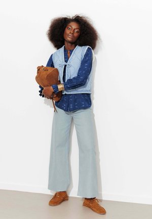 Femme aux cheveux bouclés portant un gilet bleu clair sur une chemise à motifs bleu foncé et un pantalon large bleu clair, tenant un sac en daim marron.