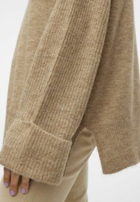 Beige gebreide trui met een textuur en ribbelpatroon, met brede manchetten, gedeeltelijk ingestopt in lichtgekleurde corduroy broek.
