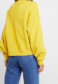 Felpa gialla oversize con maniche lunghe, polsini a coste e orlo corto, abbinata a jeans in denim blu. Texture liscia.
