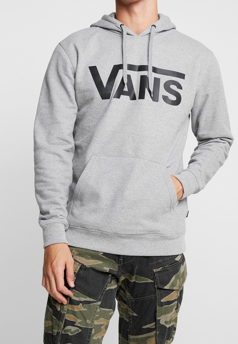 Homme portant un sweat à capuche Vans gris clair avec logo noir et pantalon cargo camouflage, main dans la poche du sweat, sur fond uni clair.