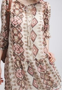 Vestido ligero y estampado con una base beige y diseños geométricos en marrón y rosa, escote redondeado y un detalle de lazo con borlas.
