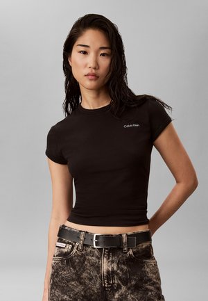 Jeune femme aux cheveux mouillés porte un t-shirt noir ajusté Calvin Klein et un jean délavé avec une ceinture noire sur un fond gris.
