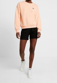 Perzikkleurige sweatshirt met ronde hals, zwarte sportbroekjes en witte hoge sneakers. Voorzien van een klein logo op de sweatshirt.