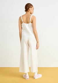 Mono blanco de lino con piernas anchas, tirantes ajustables y detalles de botones; combinado con zapatillas rosas y calcetines blancos sobre un fondo amarillo.