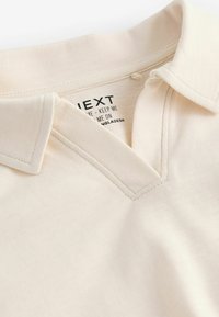 Beige polo skjorte med V-halskrave, blødt stof og synlige stikninger; inkluderer plejeetiket med mærkenavn og oprindelsesland.