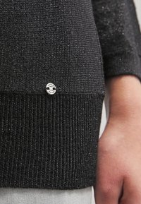 Czarny sweter o teksturowanej powierzchni z błyszczącym wykończeniem, ściągaczami na rękawach oraz małym srebrnym logo przy dolnym brzegu.