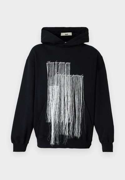 Črna kapucarica s dolgimi belimi cofki, ki visijo nad besedilom spredaj z napisom "Mir naj bo s tabo, potrpi z mano potrpežljivo."