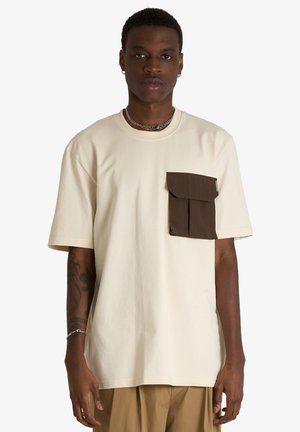 Giovane uomo che indossa una t-shirt oversize color crema con una grande tasca marrone sul petto e pantaloni beige, in piedi su sfondo bianco.