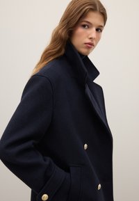 Cappotto peacoat in lana blu navy con colletto alto, design doppiopetto e bottoni dorati, caratterizzato da una texture liscia e una vestibilità sartoriale.