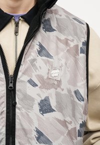 Άνδρας που φοράει γιλέκο με φερμουάρ και σχέδια πάνω από μία μπεζ μακρυμάνικη μπλούζα, με εμφανές λογότυπο «Santa Cruz» στο στήθος του γιλέκου.