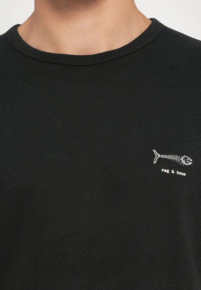 Camiseta negra de rayas con cuello redondo y un pequeño logo de esqueleto de pez blanco en el lado izquierdo del pecho, hecha de un tejido suave y texturizado.
