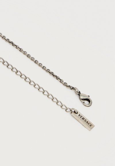 Επέκταση ασημένιου κολιέ με αλυσίδα και κούμπωμα σε σχήμα αστακού, με ορθογώνια ταμπελίτσα χαραγμένη με τη λέξη "VERSACE" σε λευκό φόντο.