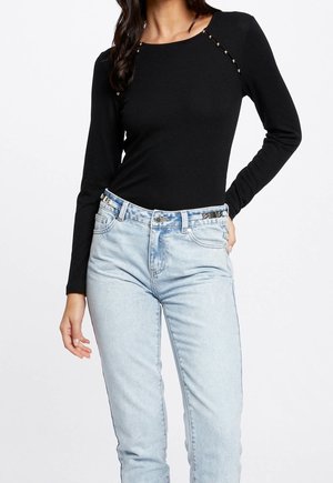 Vrouw draagt een zwart langarmig shirt met parelmoeren knopen op de schouder en lichtblauwe jeans met hoge taille, hand in de zak.