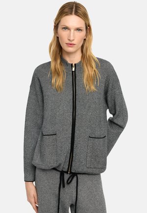 Kobieta w szarej kurtce zapinanej na zamek z czarnymi wykończeniami i dopasowanych spodniach, stojąca na jasnym, jednolitym tle.