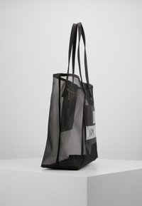 Svart mesh-handväska med dubbla handtag, som har ett dragkedja inre ficka och en djärv vit logga på ena sidan. Lätt och rymlig design.