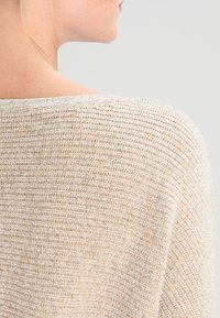 Espalda superior y cuello de una mujer que lleva un suéter de punto beige, contra un fondo claro y liso.