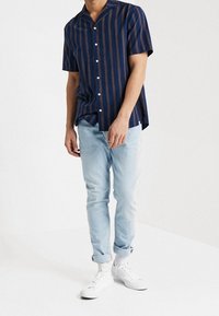 Camisa de manga corta azul marino con rayas naranjas, botones blancos y cuello chal, combinada con vaqueros azul claro y zapatillas blancas.