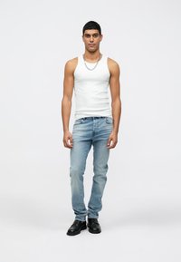 Top blanco sin mangas, jeans azul claro y zapatos negros. El modelo está de pie, con un collar de cadena plateado como acento.