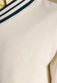 Kreminės spalvos megztinis su tamsiai mėlyna ir balta dryžuota apykakle ir subtiliu įspaustu logotipu „Joules 1989“ krūtinės srityje.