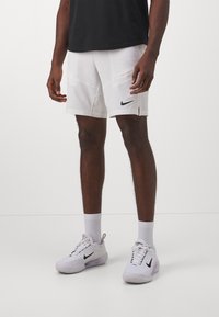 Man som bär vita Nike sportshorts, vita Nike sneakers, vita strumpor och en svart t-shirt, stående mot en enkel bakgrund.