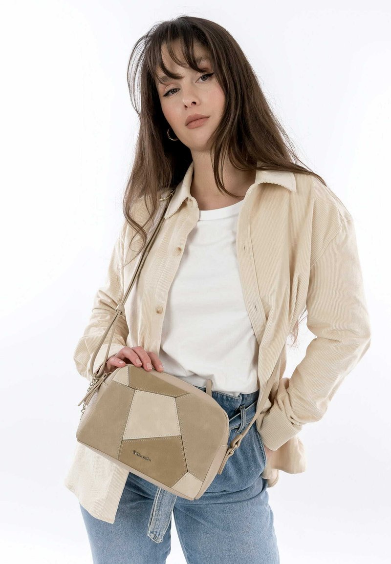 Beige geometrisk axelväska med suédepaneler och läder, med dragkedjestängning och justerbar rem, hålls av en person som bär denim.