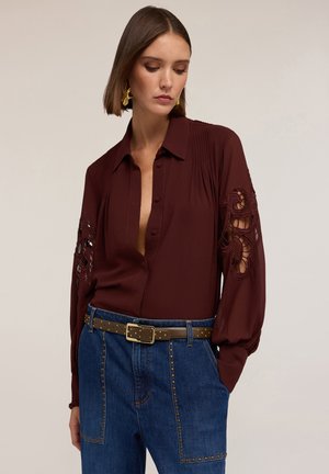 Blusa bordeaux con scollatura a V profonda, maniche lunghe con ricami intricati e intagli, abbinata a pantaloni in denim a vita alta e una cintura borchiata.