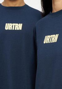 Δύο άτομα με μπλε ναυτικό πουκάμισα που φέρουν έντονο κρεμ γράμμα «URTRN» τυπωμένο στο πάνω μέρος του στήθους, φαίνονται από τον λαιμό μέχρι το μέσο του στήθους.