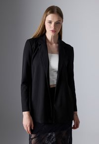 Młoda kobieta z długimi blond włosami nosi czarną marynarkę na białym topie crop oraz czarną koronkową spódnicę na neutralnym szarym tle.