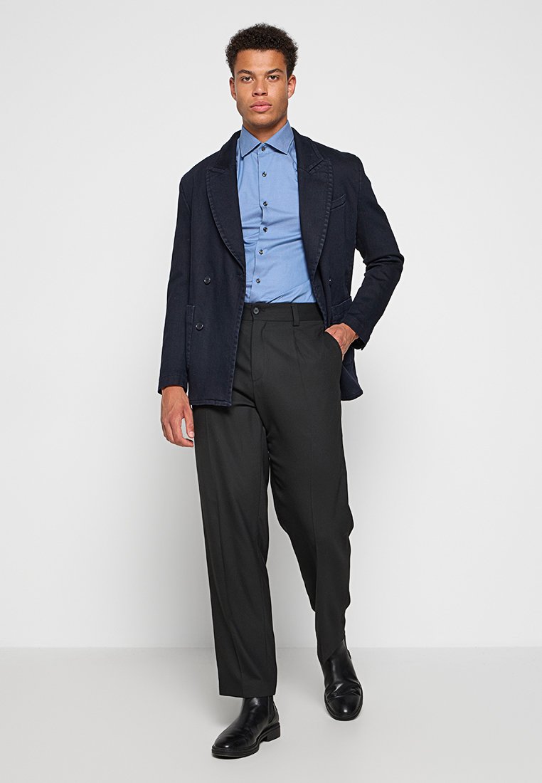 Blazer croisé bleu marine, chemise boutonnière bleu clair, pantalon noir et bottines noires, présentés sur fond blanc uni.