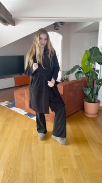 Črna halja z vezavami, ohlapen kroj, v kombinaciji s širokimi črnimi hlačami in sivimi copati. Notranji prostor z lesenim podom in rastlinami.
