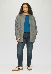 Grijze wollen mix jas met een ronde hals, voorzien van twee zijzakken en een eenvoudige knoopsluiting, gedragen over een blauwe top en donkere spijkerbroek.