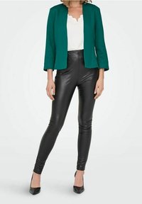 Vihreä, istuva bleiseri valkoisen blusen päällä, yhdistetty mustiin nahkahousuihin ja mustiin korkeakorkoisiin kenkiin. Sileät tekstuurit ja räätälöity siluetti.