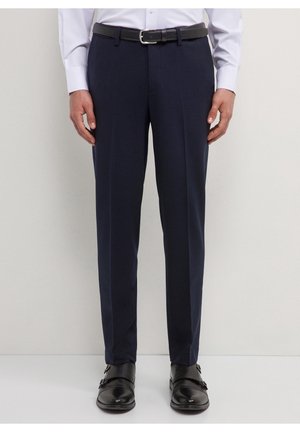 Mężczyzna ubrany w granatowe spodnie wizytowe, czarny skórzany pasek, białą koszulę i czarne eleganckie buty typu double monk strap stoi na jasnym tle.