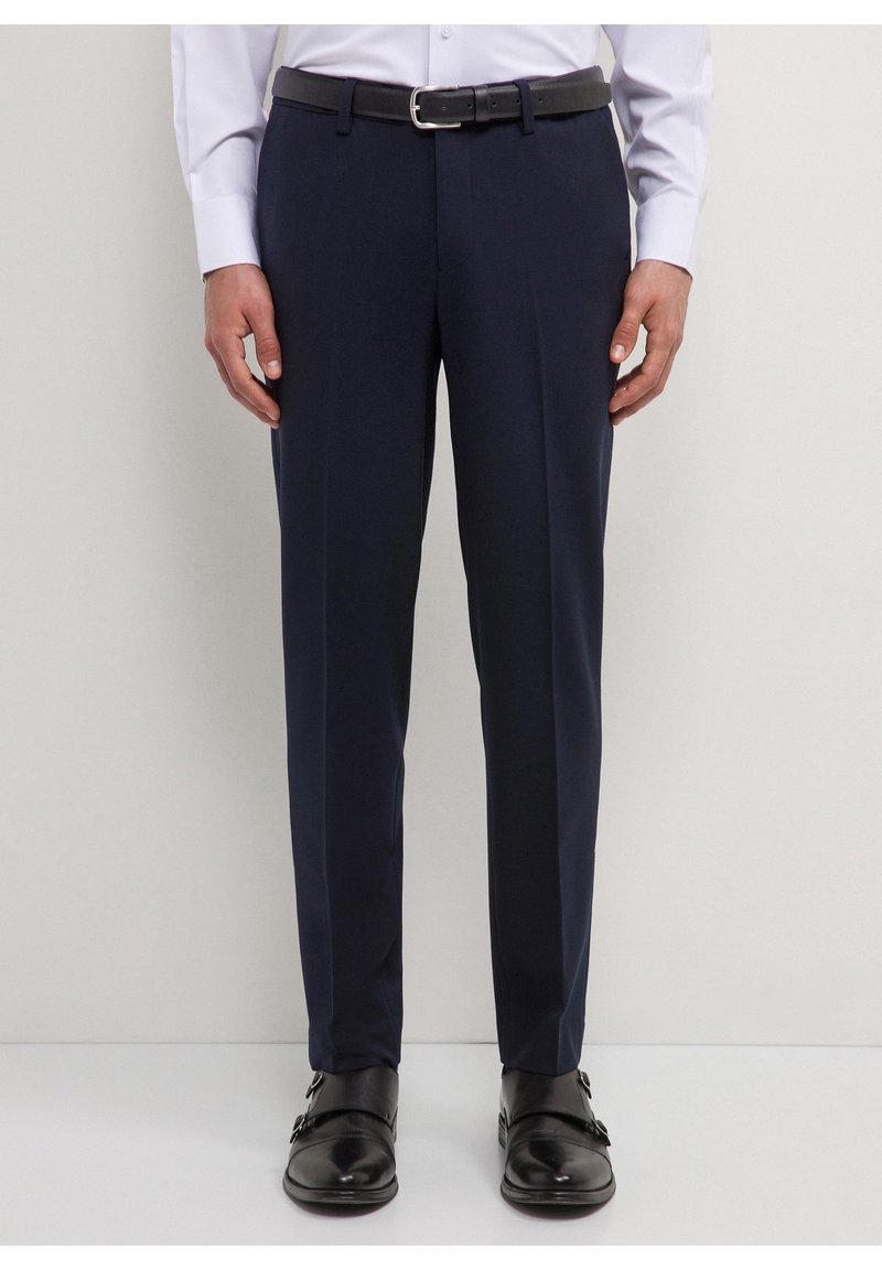 Homme portant un pantalon de costume bleu marine, une ceinture en cuir noire, une chemise blanche et des chaussures habillées à double boucle noires, debout devant un fond clair.