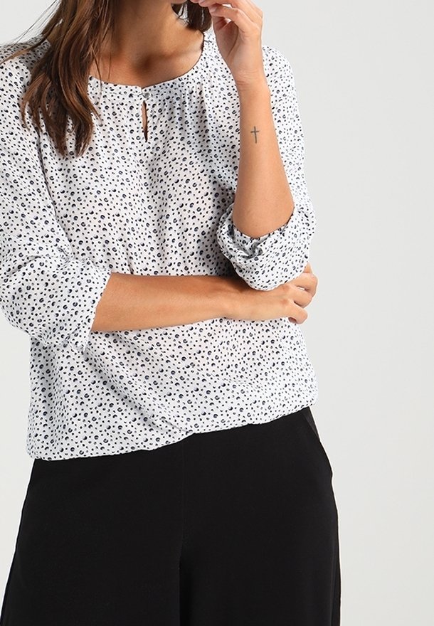 Blouse blanche à pois noirs, col rond, détail œillet, manches trois-quarts. Portée avec un pantalon noir, tissu lisse, design décontracté.