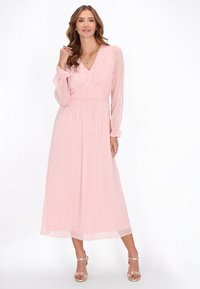 Rochie maxi din chiffon roz deschis cu decolteu în V, mâneci lungi, talie strânsă și un model subtil cu puncte aurii. Asortată cu sandale metalice.